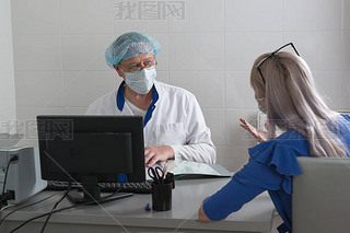 A doctor in a white coat, mask and cap in the office sits at a table with a patient and listens to h