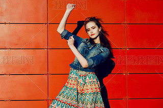 Young teenager girl hing fun, posing and iling near red wall background in skirt and jeans jacke