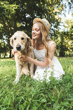 美丽的年轻女孩在白色礼服和草帽抚摸金毛猎犬，而微笑，坐在草地上，看着狗