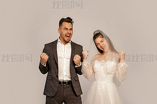 excited newlyweds showing win gesture while looking at camera isolated on grey