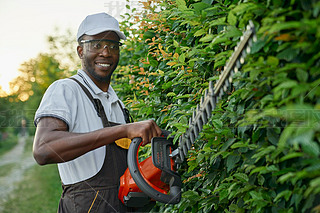 Smiling afro gardener using hedge trimmer for cutting bushes