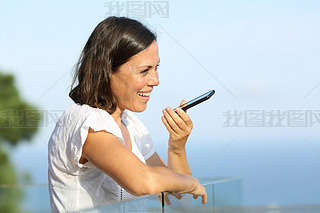 Happy adult woman using voice recognition on art phone standing in a balcony on the beach