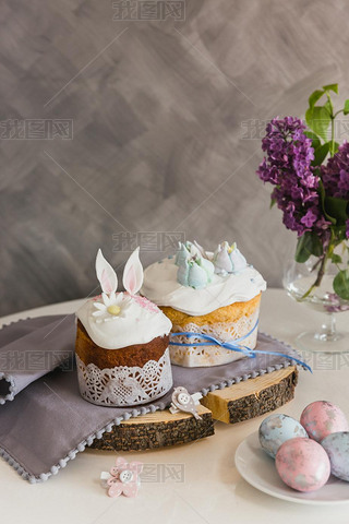 Easter Cakes on wooden decorated with rabbit ears, eggs on whith plate on foreground, lilac on backg