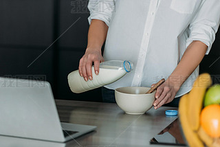 cropped view of woman pouring milk into bowl near laptop in kitchen