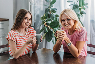 happy mother and daughter iling at camera while sitting at kitchen table with tea cups