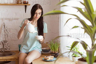 Beautiful brunette woman sitting in the kitchen, on the table, drinking tea and eating waffle, desse