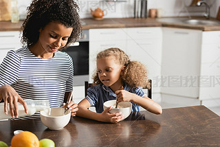 young african american woman in striped t-shirt pouring milk into bowl during breakfast with daughte