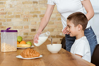 Mother pouring milk in bowl with cereals near iling son at kitchen table 