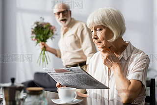 Selective focus of senior woman reading newspaper near positive husband with bouquet 