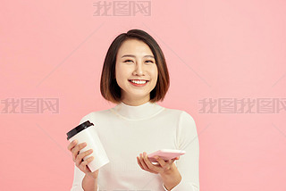 Close up portrait of young Asian woman holding coffee cup and artphone over pink background.