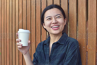 Smiling young lady standing at the wooden wall outdoors and holding a cup of coffee.