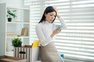 Upset young brunette female sitting on her desk, holding mobile phone in one hand and touching her h