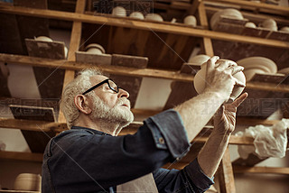 Senior potter in apron and eyeglasses examining ceramic bowl at workshop