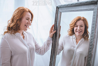 beautiful iling mature woman standing near mirror and looking at reflection