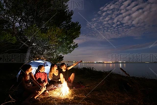 一群四游客坐在海边的篝火前的旅游帐篷树下, 胡子男子指着明亮的蓝色傍晚的天空, 在清澈的水背景。旅游业和野营概念.