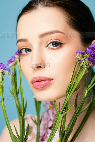 young woman looking at camera near blooming flowers isolated on blue 