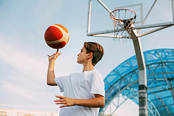 一位身穿白色T恤的少年篮球运动员站在篮球场上，用手指捻着篮球。体育与健康生活方式的概念