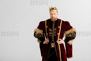 handsome king with crown with hands on hips looking at camera isolated on grey