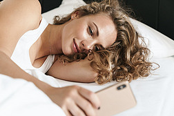 靠近一个年轻美丽的金发女人，她用手机躺在床上的枕头上，面带微笑