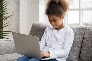 Curious preschool african mixed race girl using laptop on couch, little art black kid typing on co