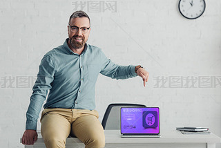 handsome businesan sitting on table and pointing with finger at laptop with web-shopping website 