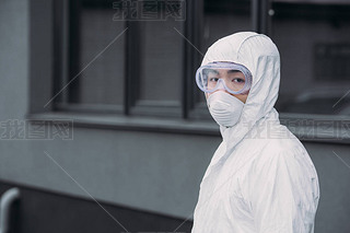 asian epidemiologist in hazmat suit and respirator mask looking at camera while standing on street