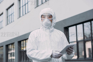 asian epidemiologist in hazmat suit and respirator mask holding digital tablet and looking at camera