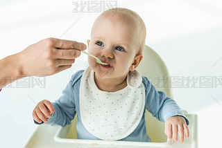 Mother with spoon feeding cute baby boy on feeding chair on white background