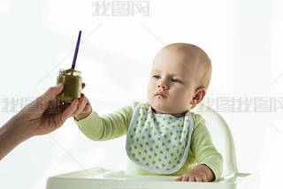 Selective focus of mother giving jar of baby nutrition to infant on feeding chair on white backgroun