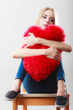 破碎的心的爱的概念。悲伤不快乐的女人坐在椅子上拥抱红色心形枕头