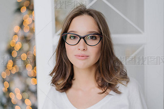 Headshot of lovely young female model in transparent glasses, has dark hair, dressed in casual cloth