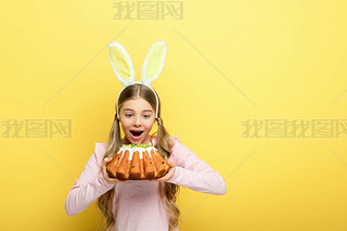 shocked kid with bunny ears holding easter cake isolated on yellow 