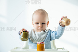 Selective focus of baby holding jars of vegetable baby nutrition while sitting on feeding chair on w