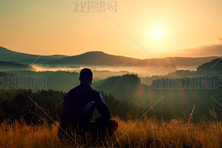 Hiker in squatting position in high grass meadow  enjoy the colorful sunrise scenery