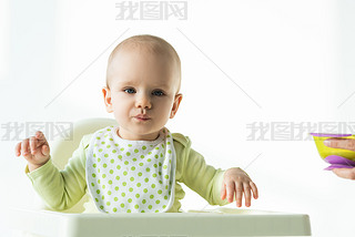 Adorable baby looking at camera while sitting on feeding chair near mother with bowl of baby nutriti