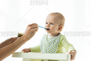 Mother with jar of vegetable baby nutrition and spoon feeding infant on feeding chair isolated on wh